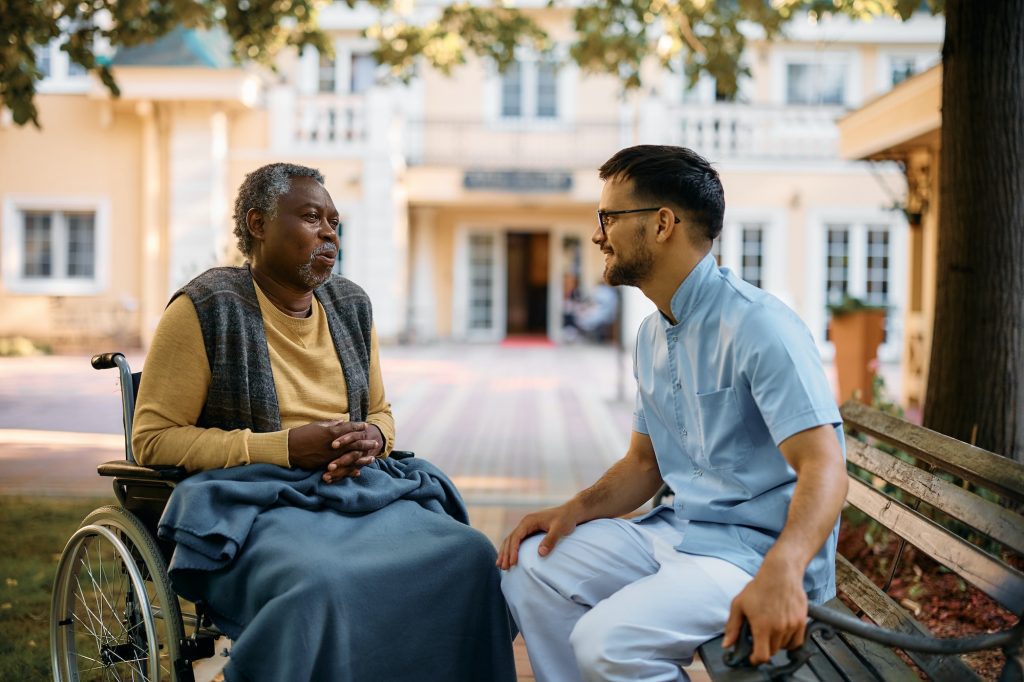 Black senior in wheelchair talking to male nurse while relaxing in park of residential care home.