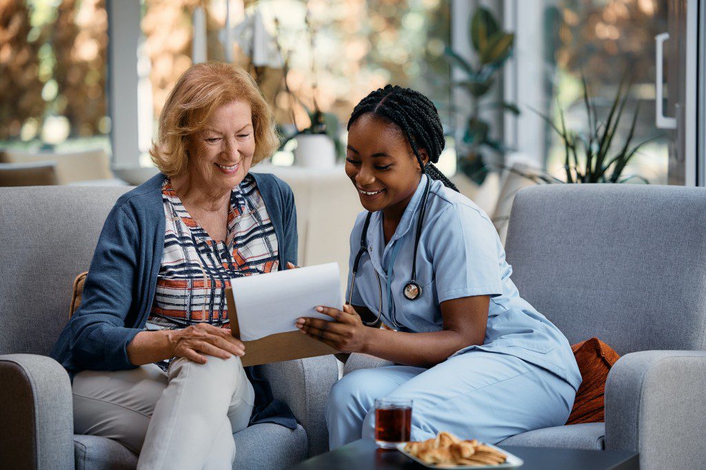 Happy senior woman and black female caregiver analyzing medical data during home visit.