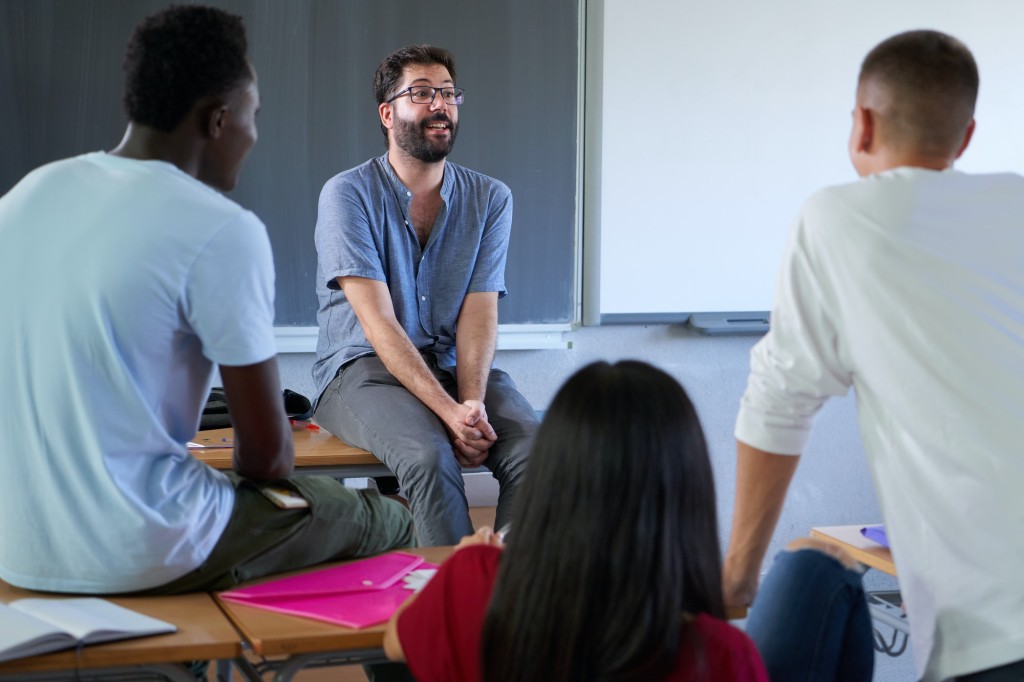 Teacher involved with students at college sitting on his desk while giving an interactive class.