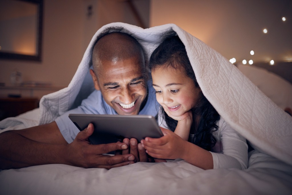 Father, kid and smile with tablet at night under blanket of online games, reading ebook or storytel