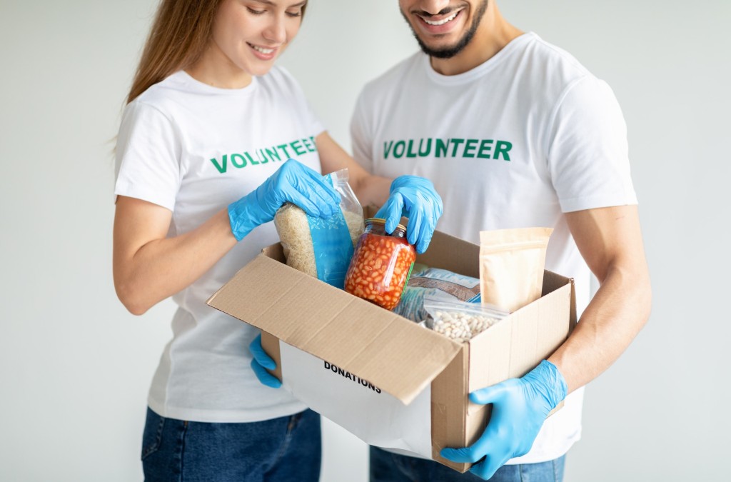 Young volunteers packing food donations in cardboard box standing over light background, crop