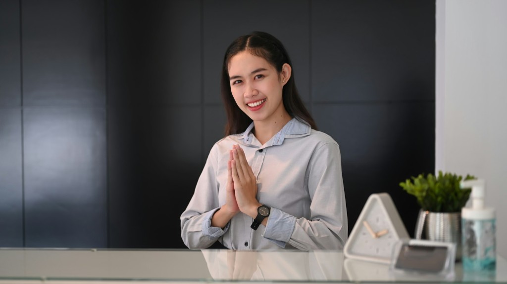 Young asian woman receptionist sitting at reception desk and raising her hands pay respect.