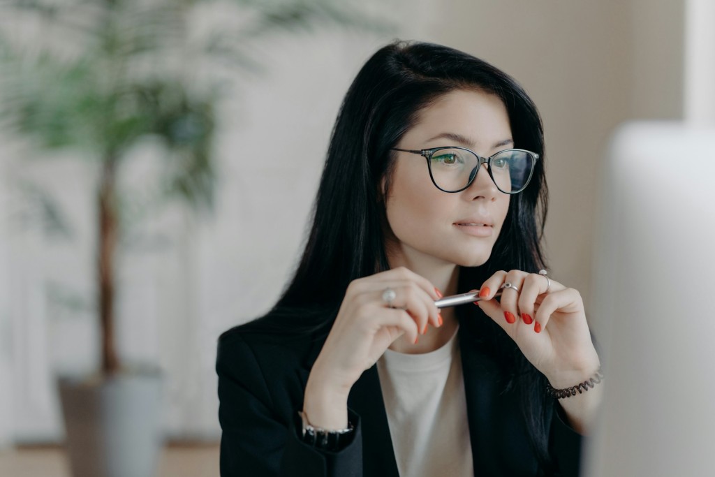 Skilled female freelancer concentrated at computer display, wears optical glasses, holds pen